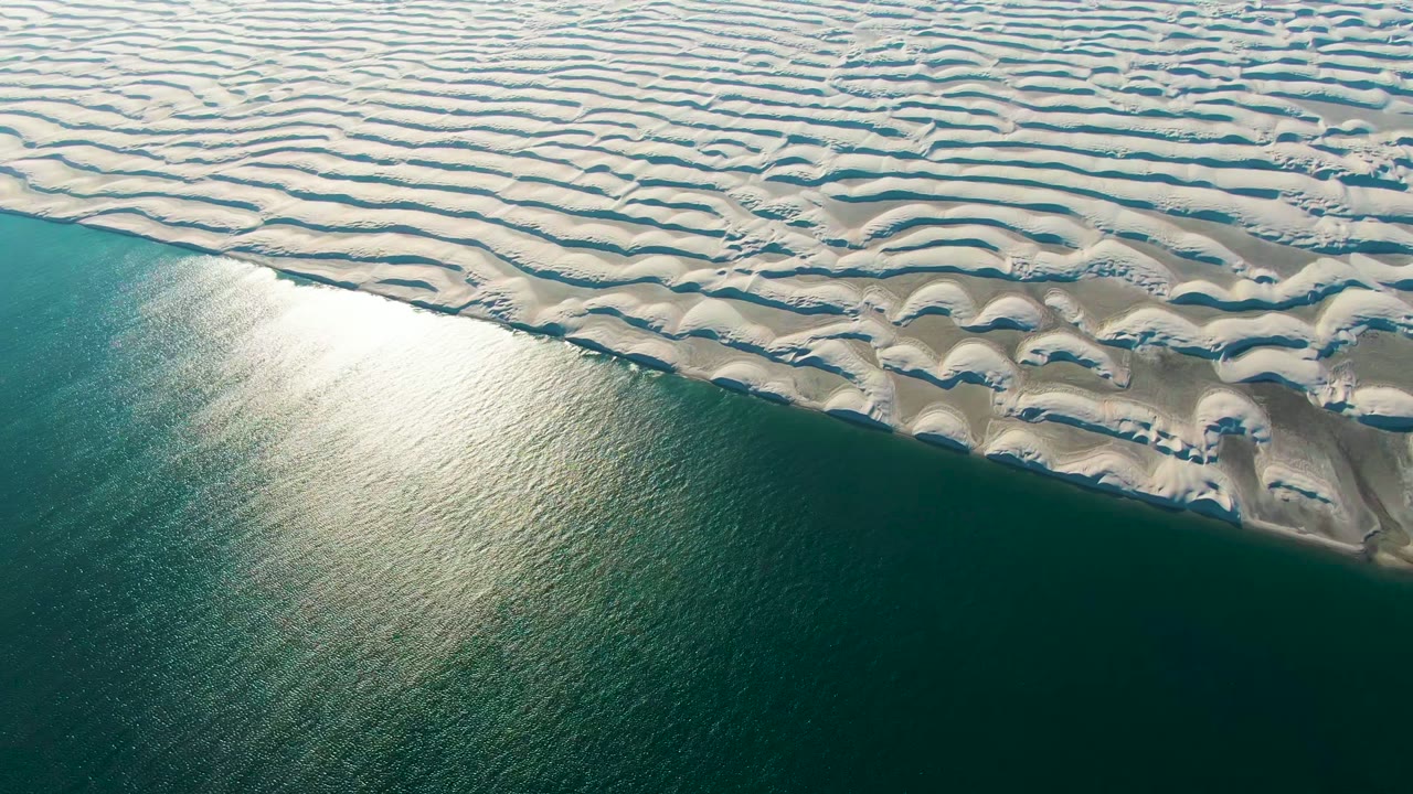 Aerial view in Guerrero Negro, Mexico, with repeating white dune ridges in the upper area, a narrow irregular shore edge, dark green ocean below, and no visible people.