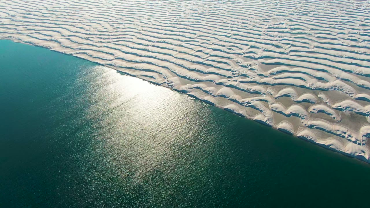 At 0:00, white rippled sand dunes meet teal ocean water in Guerrero Negro, Baja California Sur, with a bright sun reflection near the shore and no visible people.