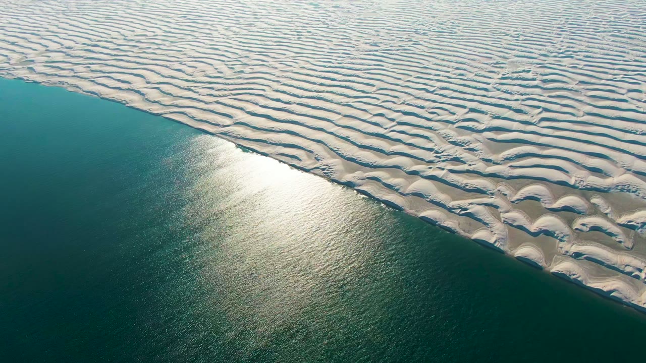 At 0:02, a bright patch of sunlight spreads across the calm ocean below the white dune field in Guerrero Negro, with no visible people.