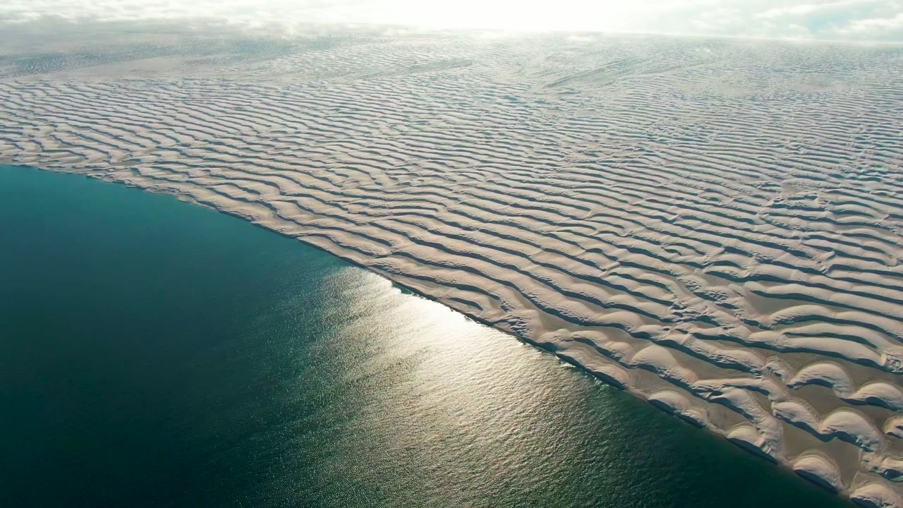 Aerial view over Guerrero Negro, Baja California Sur, with rippled pale sand dunes meeting dark teal ocean water, bright sunrise glare on the surface, and no visible people.