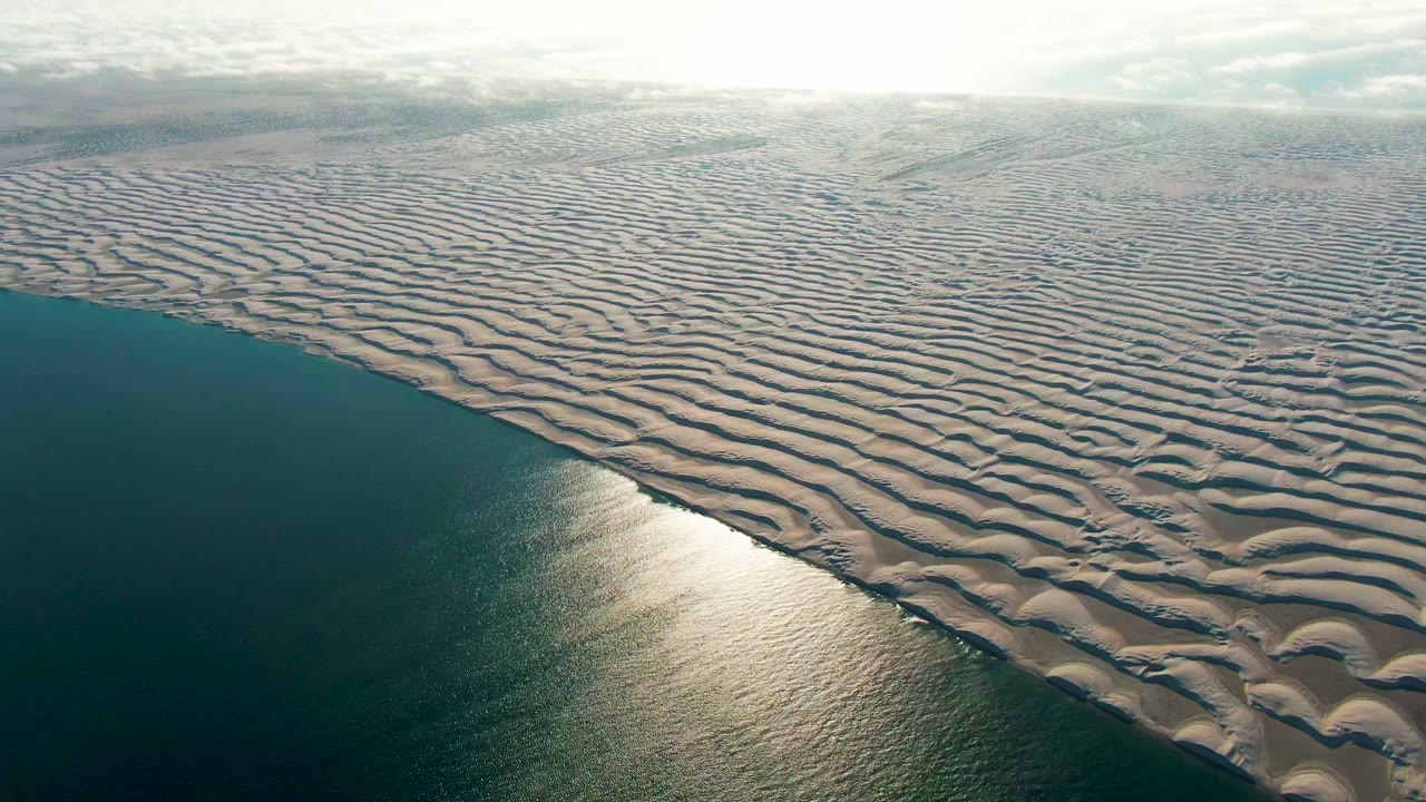 Rippled coastal dunes border the ocean with bright reflected sunlight on the water.