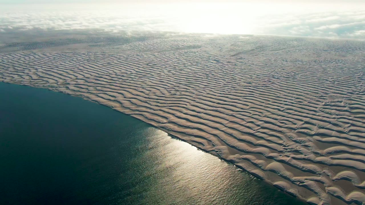 The Guerrero Negro coastline remains in aerial view, with repeating sand ridges covering most of the frame, dark ocean at left, hazy cloud bands, and no visible people.