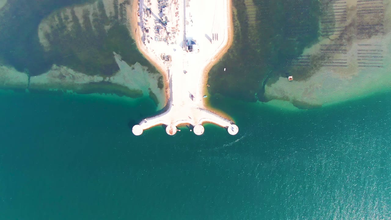 At 0:00, an overhead aerial view in Guerrero Negro shows a pale pier at the top of frame, turquoise water below, shallow sand and rock patches on both sides, and no visible people.