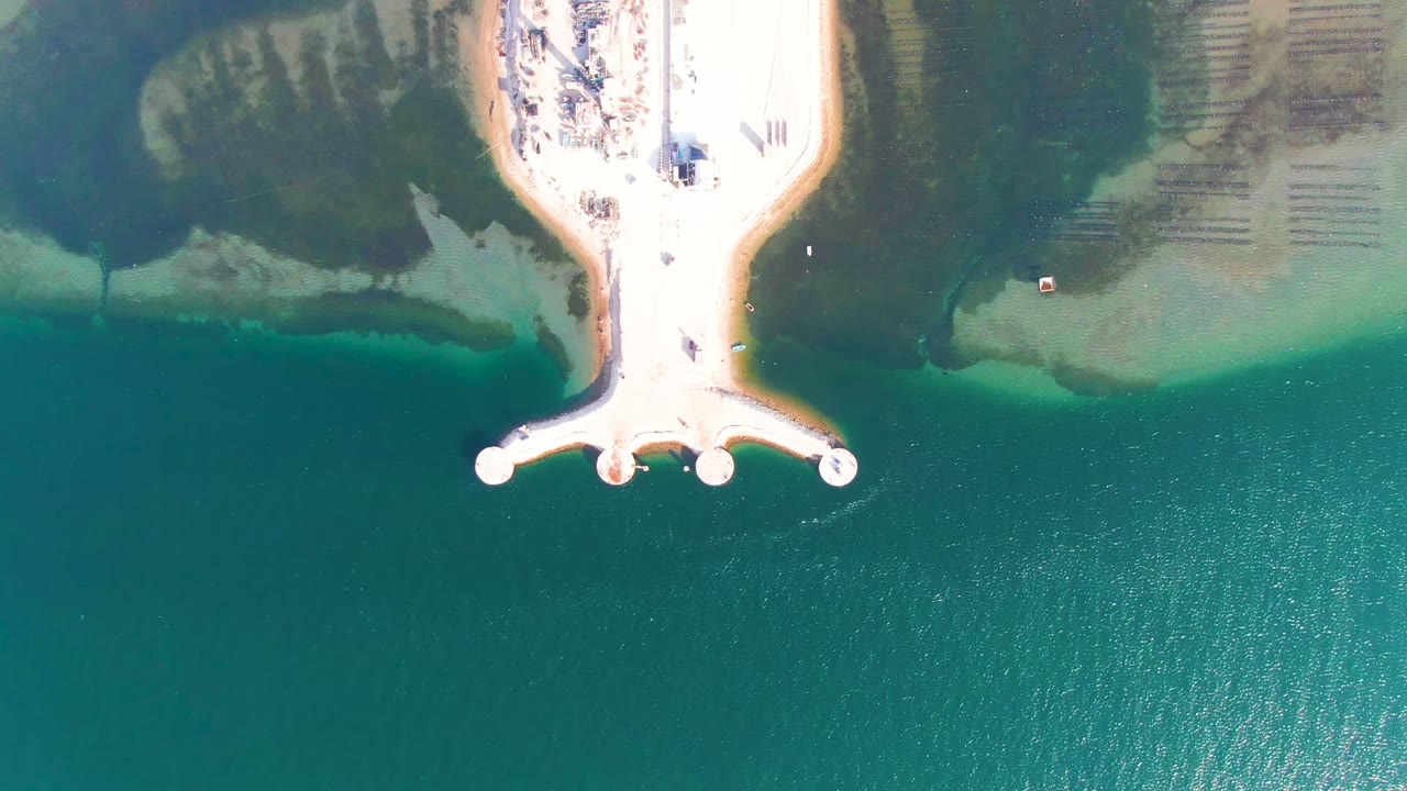 At 0:02, the aerial view shows the pale pier against darker submerged shoreline patches and lighter sandy flats in Guerrero Negro, with open water below and no visible people.