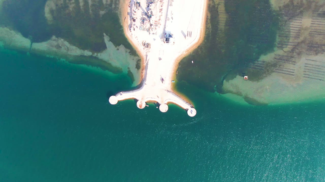 Overhead frame from Guerrero Negro, Baja California Sur, showing a bright sandy pier reaching into turquoise water with dark shallow shoreline areas and tiny people visible near the beach.