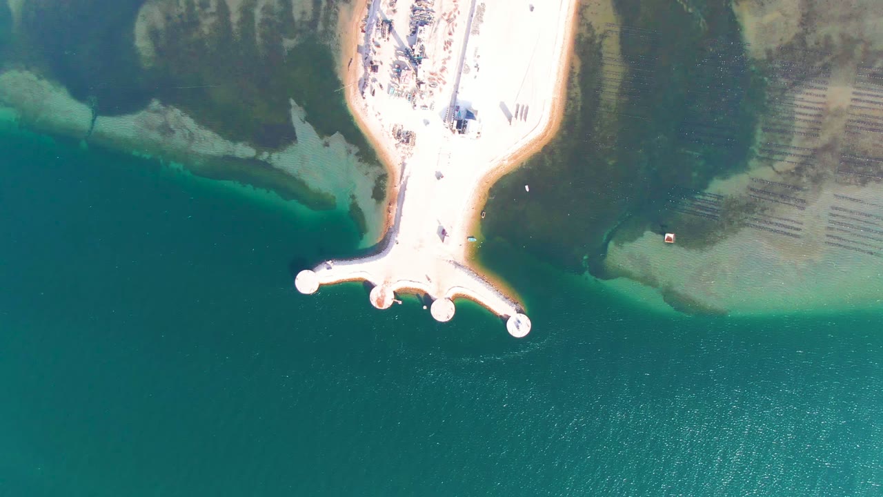 Top-down Guerrero Negro view with the pier at the upper center, turquoise water filling the lower half, green shoreline vegetation at the top, and tiny people visible on the bright sand.