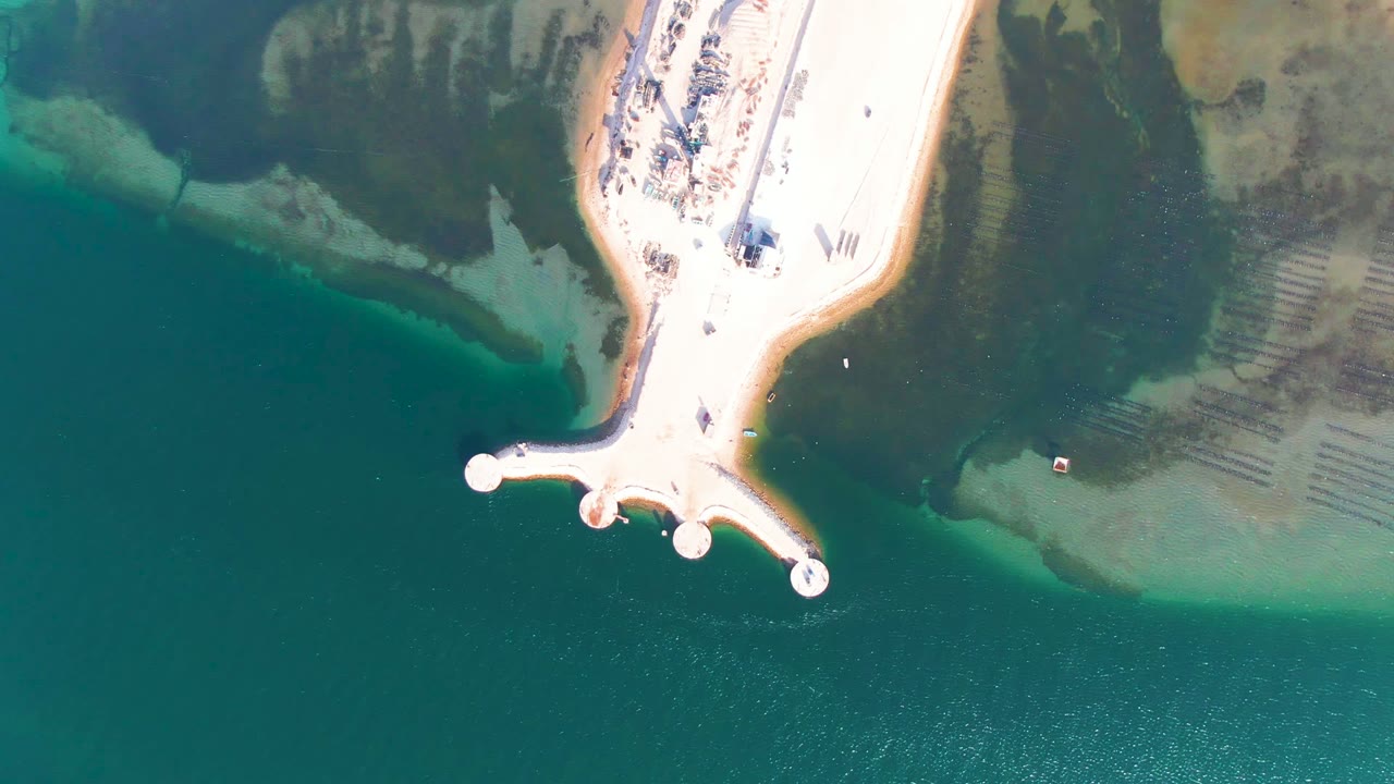 Overhead frame from Guerrero Negro, Baja California Sur, showing a pale sandy pier with round ends extending into turquoise and dark green water, with no visible people.