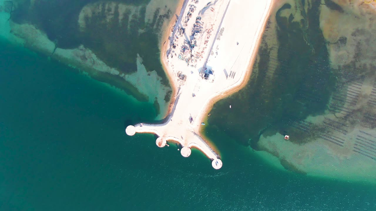 Drone-style overhead frame from Guerrero Negro, Baja California Sur, showing the pale pier angled from the upper right toward round platforms above darker green water, with no visible people.