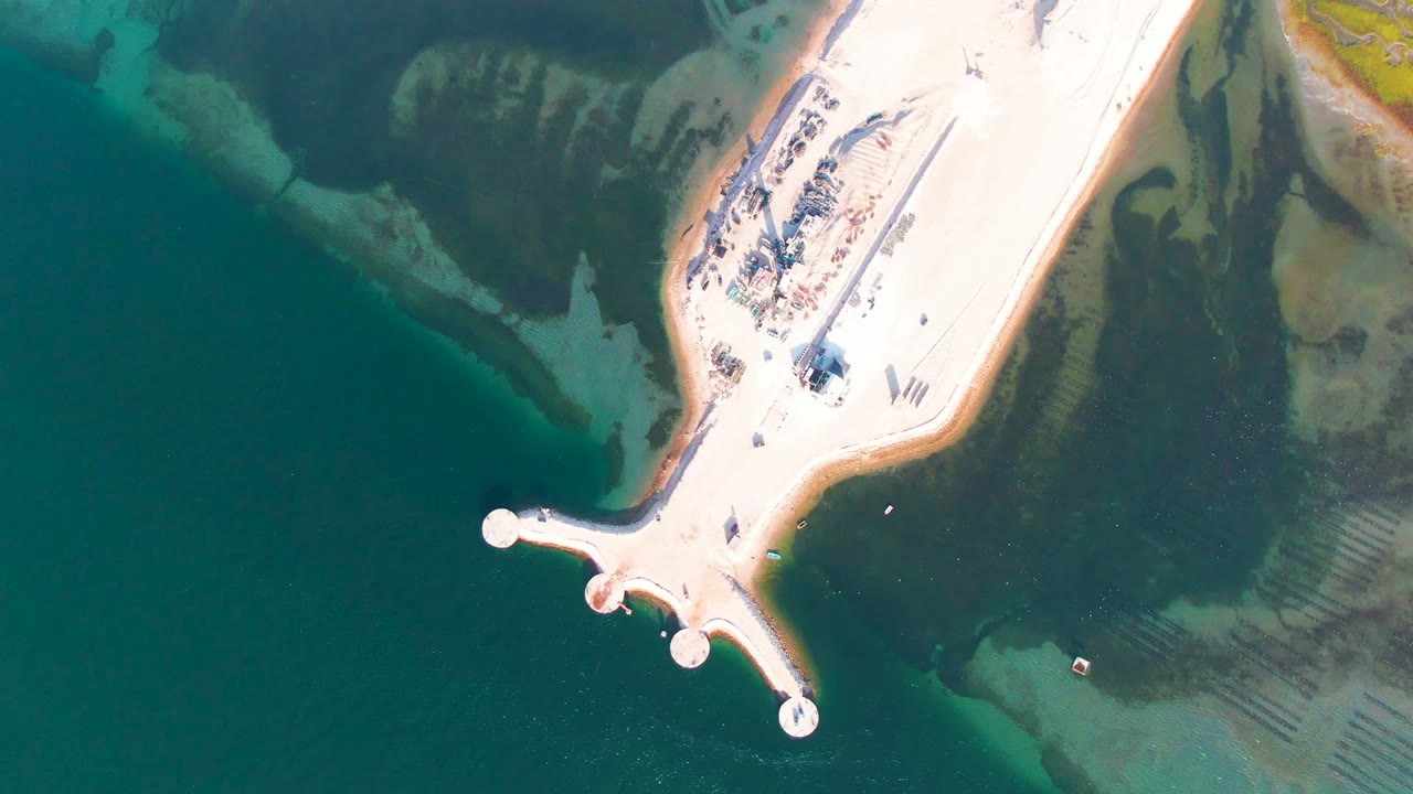 Overhead frame from Guerrero Negro, Baja California Sur, showing a pale sandy causeway with rounded pier ends extending into blue green water, with no visible people.
