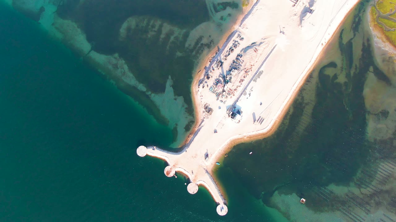 At 0:02, the overhead Guerrero Negro frame keeps the causeway centered between turquoise water on the left and darker shallow water beside the sandy shore, with no visible people.