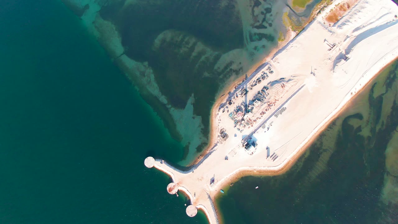 At 0:02, a top-down frame shows the rounded road junction near the lower center, dark water on the left, pale exposed ground on the right, and no visible people in the Guerrero Negro scene.
