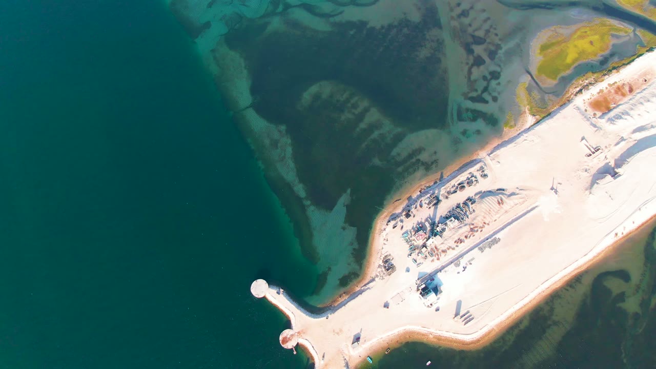 Top-down aerial frame over Guerrero Negro, Baja California Sur, showing a pale coastal causeway, small shore structures, dark teal water, green shallows, muddy flats, and no visible people.