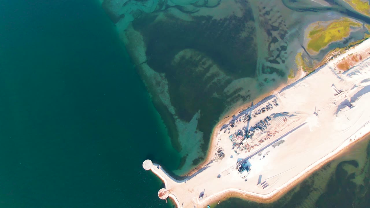 A second aerial frame over Guerrero Negro keeps the pale causeway along the lower right, with dark open water to the left, mottled shallow flats above, and no visible people.