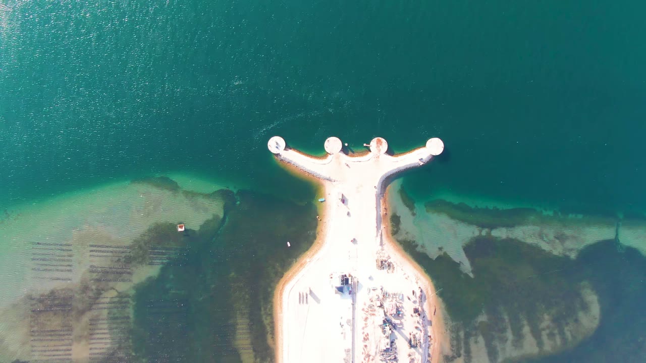 Overhead frame from Guerrero Negro, Baja California Sur, showing a bright T-shaped pier extending into clear shallow water with darker seabed patches on both sides and tiny people visible on the platform.
