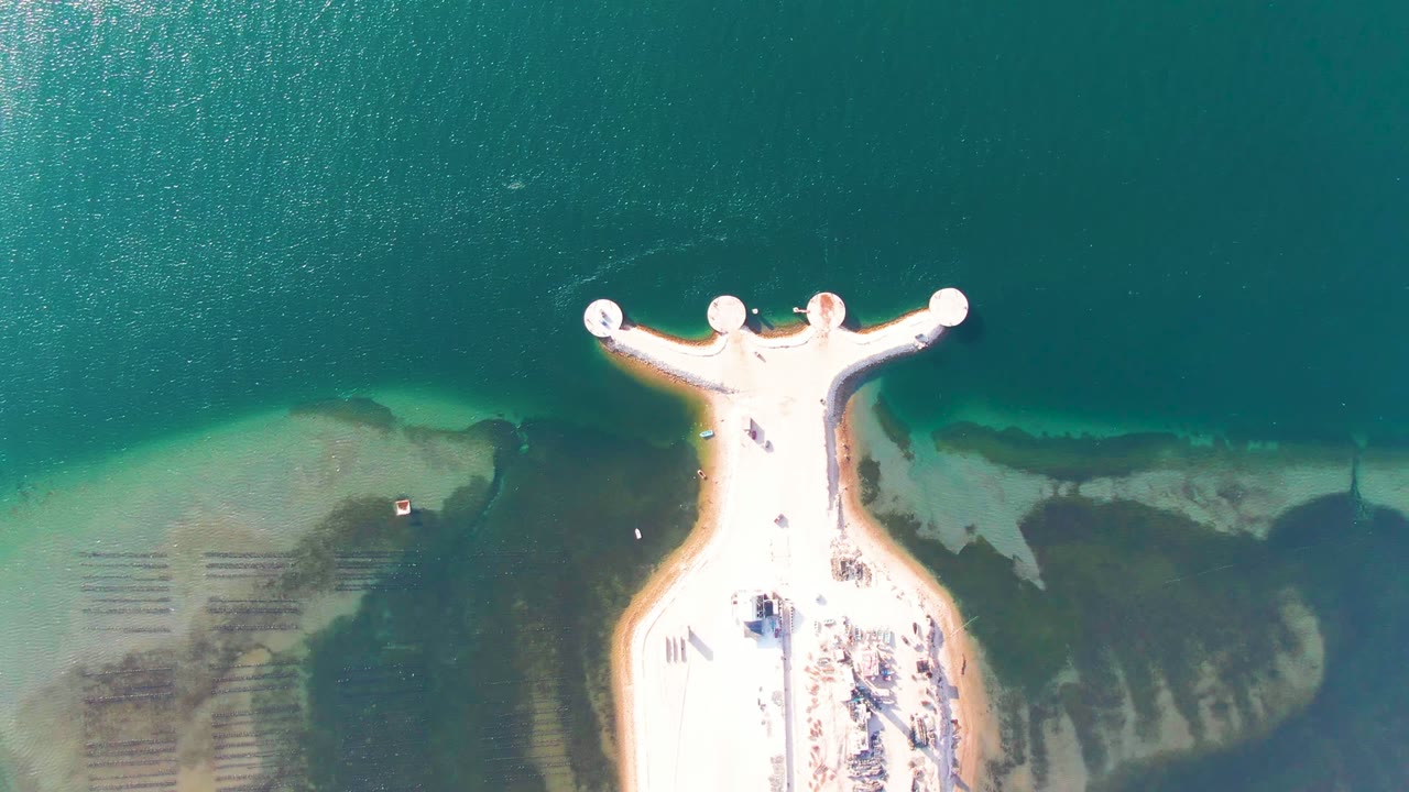 Vertical aerial frame from Guerrero Negro showing the bright pier against blue green ocean, with darker underwater patches to the right, pale sand to the left, and people visible as small dots on the pier.