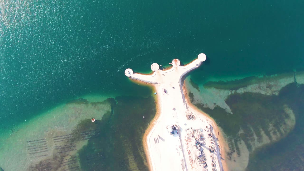 Opening overhead frame in Guerrero Negro, Baja California Sur, Mexico, with a pale pier reaching into blue-green water, shallow sand and rock shapes along the shore, and no visible people.
