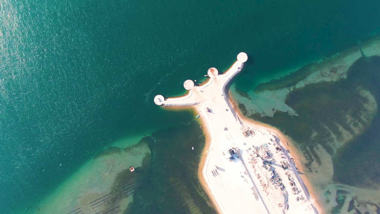 Overhead Guerrero Negro view with the pier tip near the upper frame, round platforms over green-blue water, sandy edge, and no visible people.