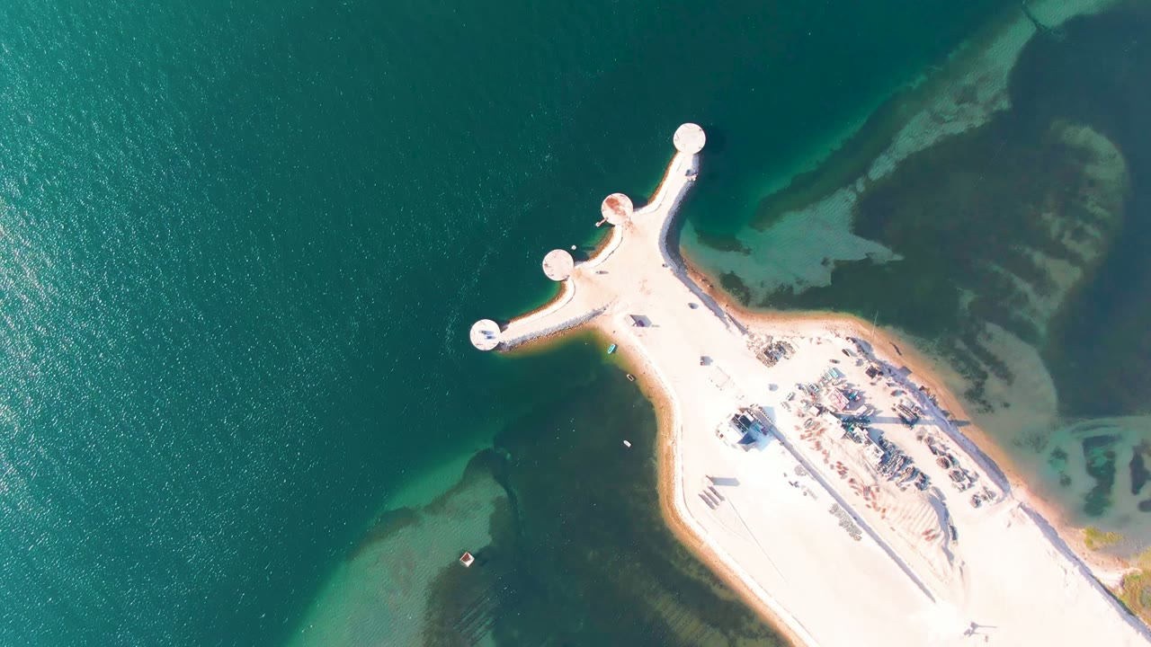 Opening aerial frame over Guerrero Negro, Baja California Sur, with a pale sandy pier projecting into green-blue water and no visible people.