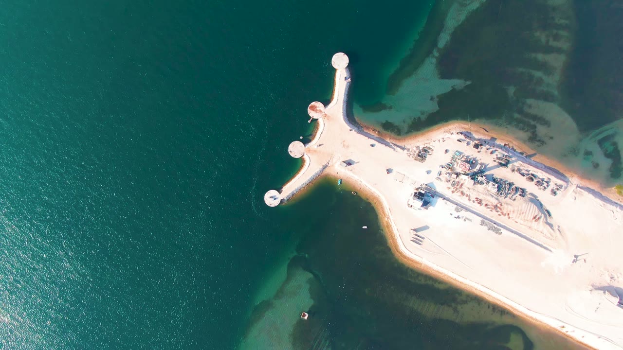 Overhead frame from Guerrero Negro, Baja California Sur, Mexico, showing a pale pier with circular platforms reaching into green-blue water and darker tidal flats, with no visible people.