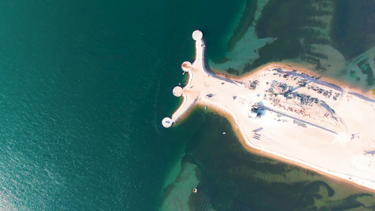 Top-down frame from Guerrero Negro, Baja California Sur, Mexico, showing the pale pier at the edge of exposed sandy land, surrounded by green water and darker shallows, with no visible people.
