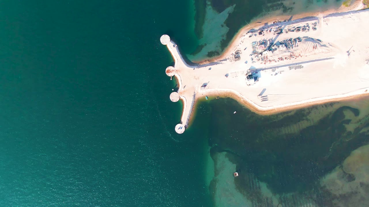 Top-down Guerrero Negro frame with the pier projecting from a bright sandy shore into teal water and darker shallow areas near the lower right, with no visible people.
