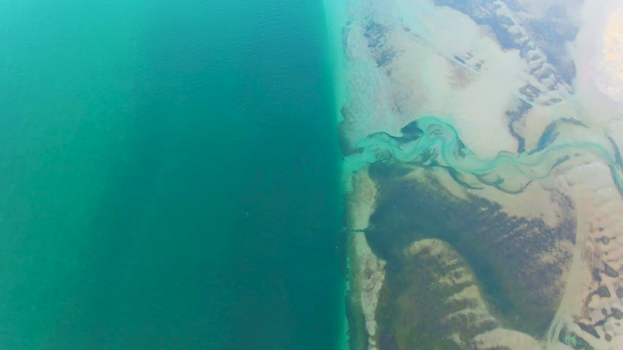 Overhead view in Guerrero Negro, Baja California Sur, with deep turquoise ocean on the left and a pale winding river channel meeting sandy shoreline on the right, no visible people.