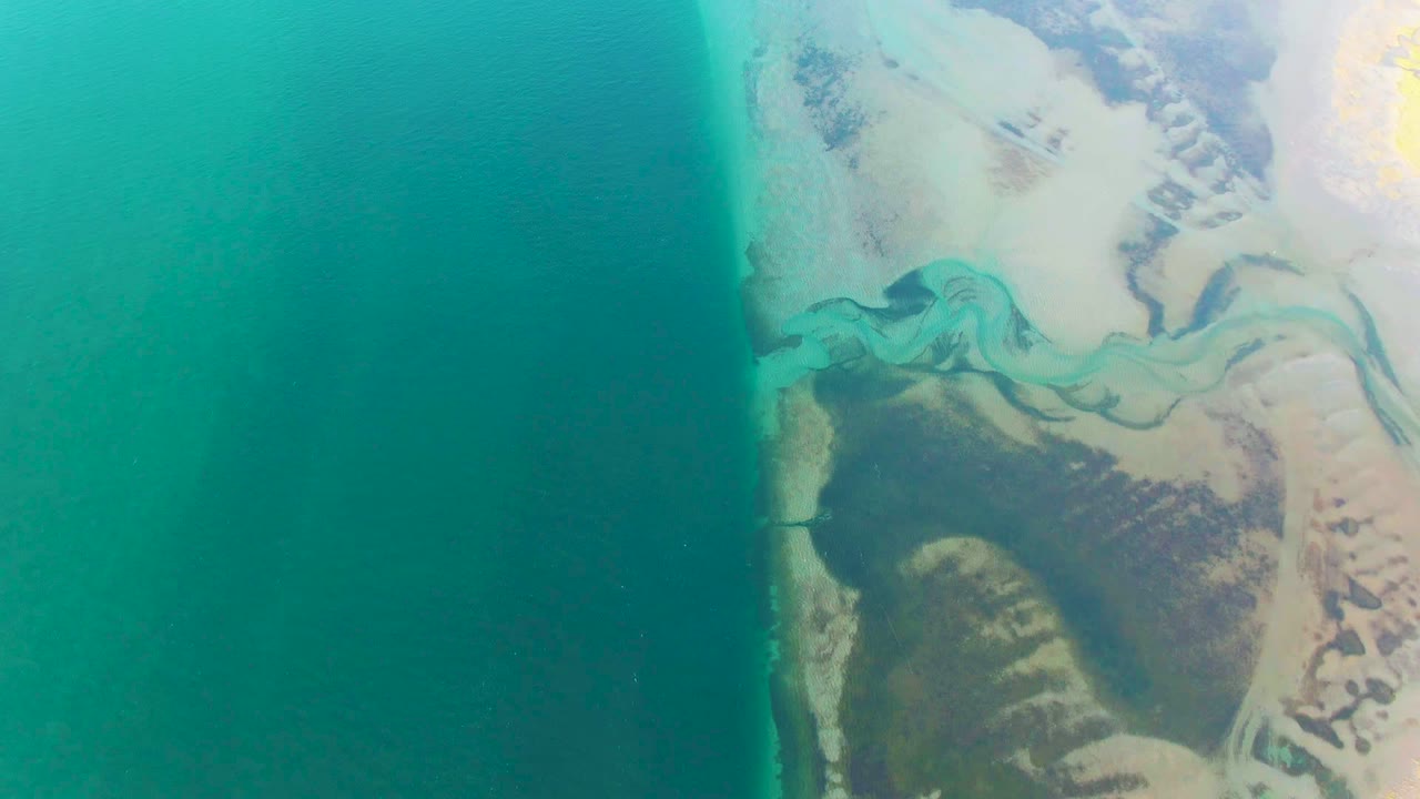 Overhead Guerrero Negro coastal view with broad turquoise ocean on the left and branching pale river water crossing sandy terrain on the right, no visible people.