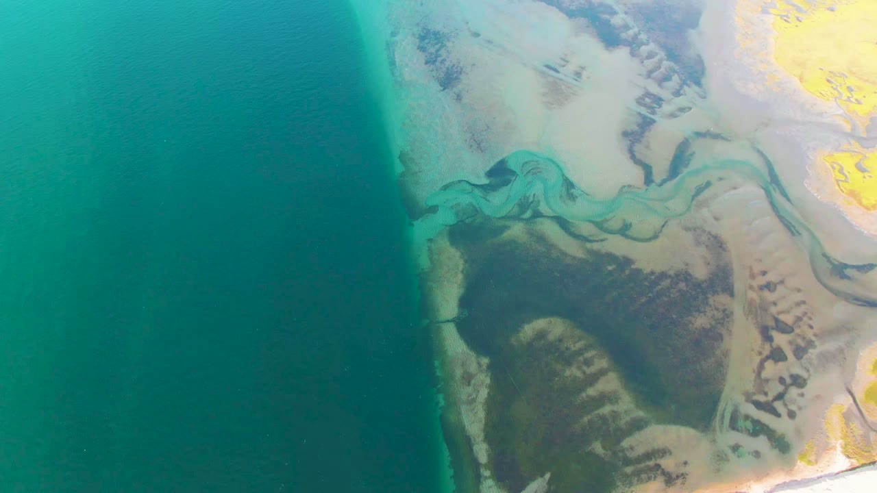 At 0:00, an aerial view over Guerrero Negro shows teal ocean on the left, pale sand and green-blue delta channels on the right, darker vegetation near shore, and no visible people.
