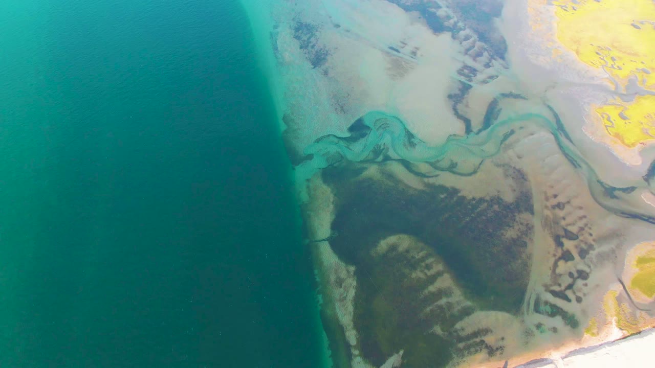 At 0:02, the Guerrero Negro coast remains framed from above, with the ocean as a broad teal field and the green-blue delta channels more centered against pale sand and vegetation, with no visible people.