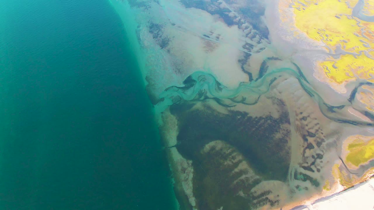 Opening aerial frame over Guerrero Negro, Baja California Sur, with dark blue ocean on the left, pale tidal flats and green channels through the center, yellow land at upper right, and no visible people.