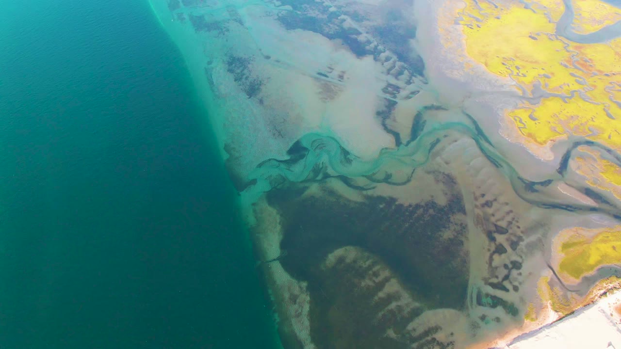 At 0:02, the aerial frame shows turquoise shallows and dark channel lines cutting through pale coastal flats near Guerrero Negro, with yellow land at the upper right and no visible people.