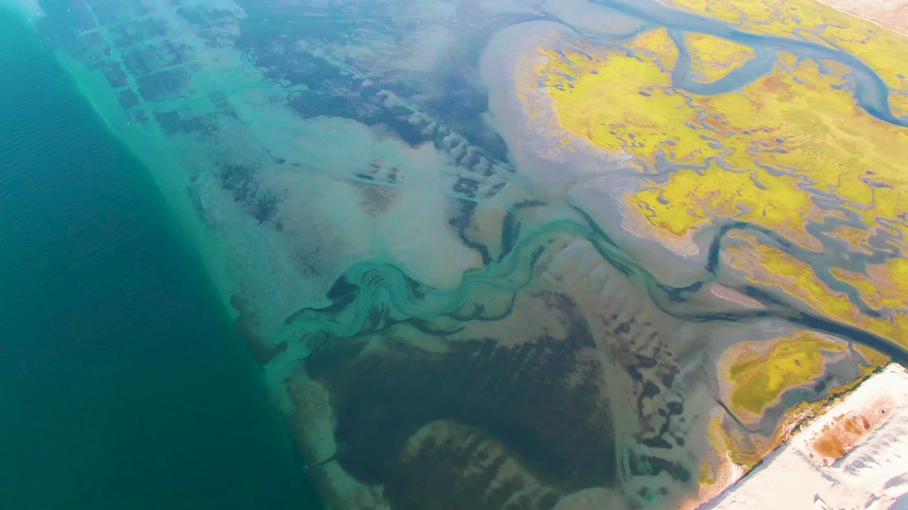 Overhead frame from Guerrero Negro, Baja California Sur, where shallow turquoise water meets dark mudflats, pale sand, yellow exposed land, and no visible people.