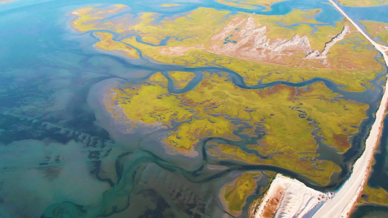 Opening aerial frame over Guerrero Negro wetland flats, with turquoise water on the left, green and yellow delta terrain, dark winding channels, a pale road at right, and no visible people.