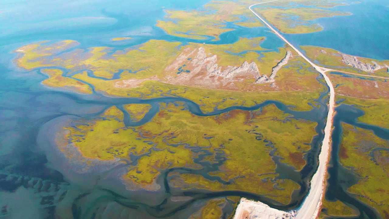 Drone view of a pale road dividing green marsh and blue channels.