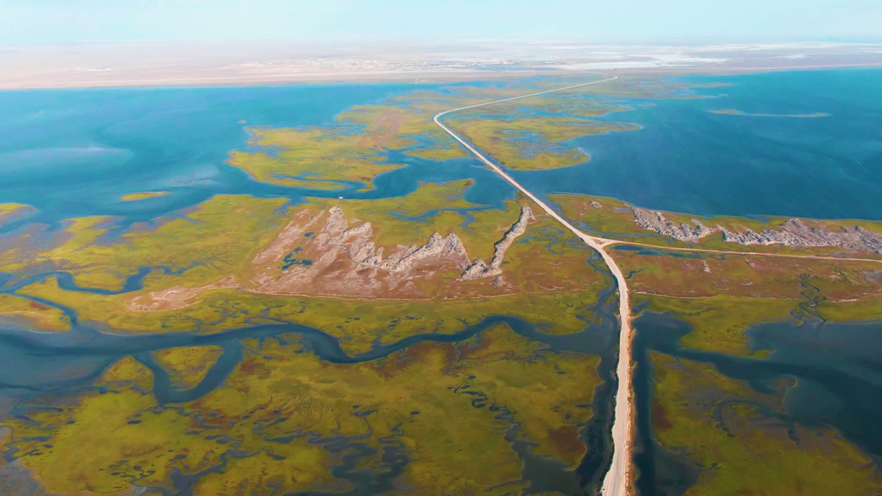 Aerial frame over Guerrero Negro, Baja California Sur, with a pale road cutting through green marshland, blue water channels, tan rocky hills, a flat horizon, and no visible people.