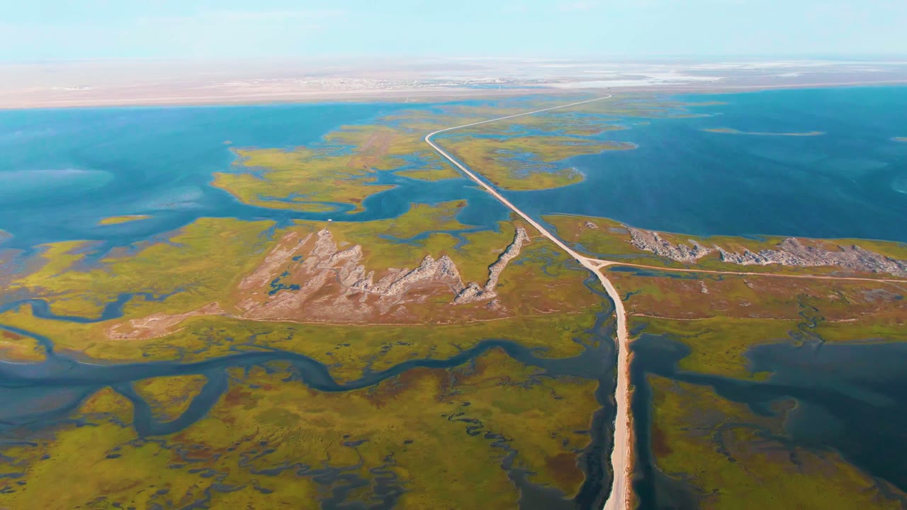 High aerial frame over Guerrero Negro, Baja California Sur, with the road leading toward broad blue water, green wetland islands on both sides, tan ridge lines, and no visible people.