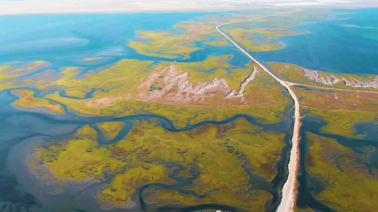 Drone view of marsh channels, a long road, rocky ground, and open blue water.