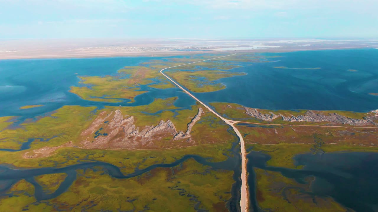 A wide aerial frame over Guerrero Negro shows a pale dirt road running through green wetland channels toward blue open water and low rocky outcrops, with no visible people.