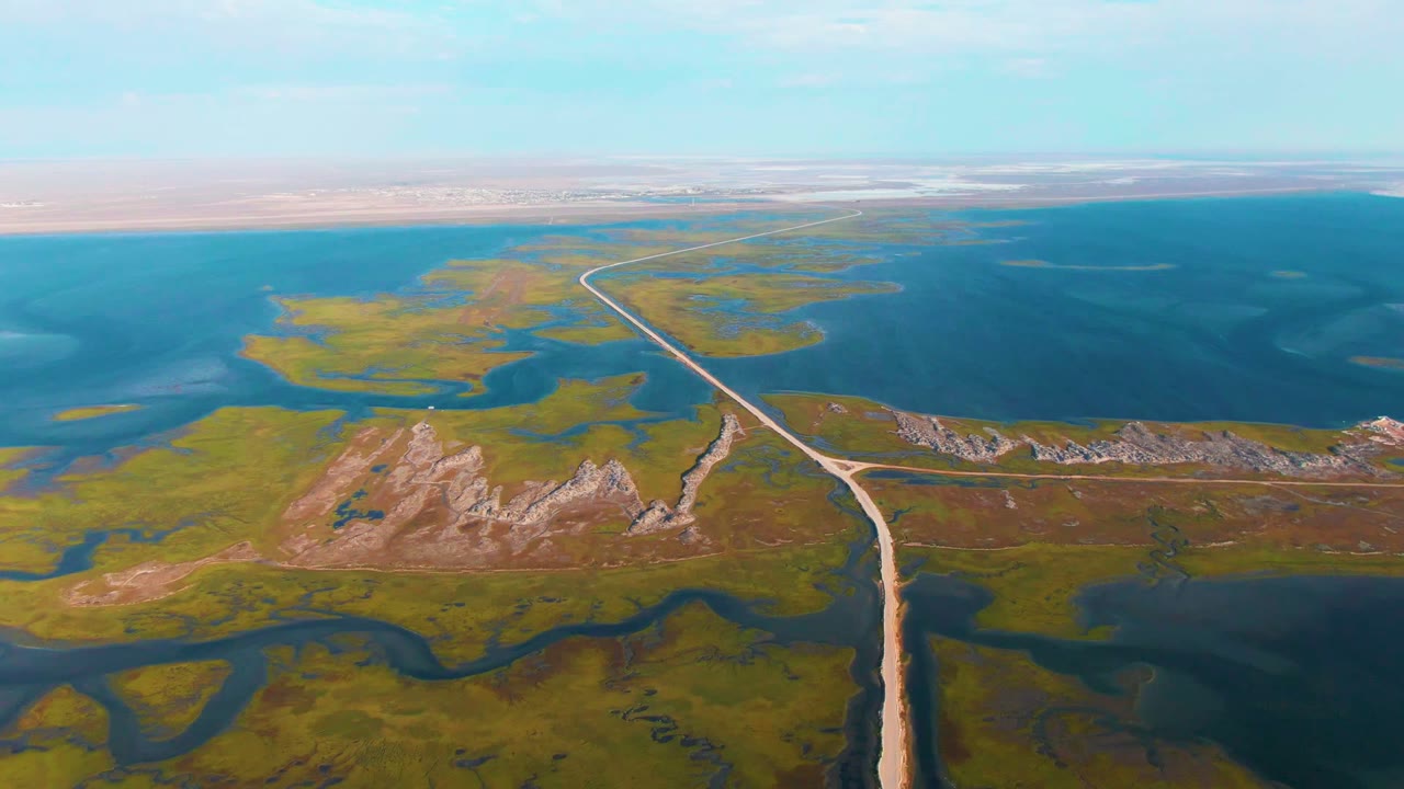 Dirt road curves through marshland beside a large blue water body.