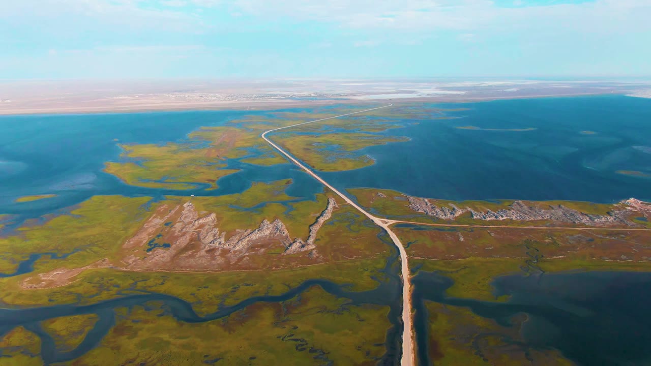 The frame looks down over Guerrero Negro wetlands where a pale road crosses green marsh and shallow water toward a line of low rocky ridges, with no visible people.