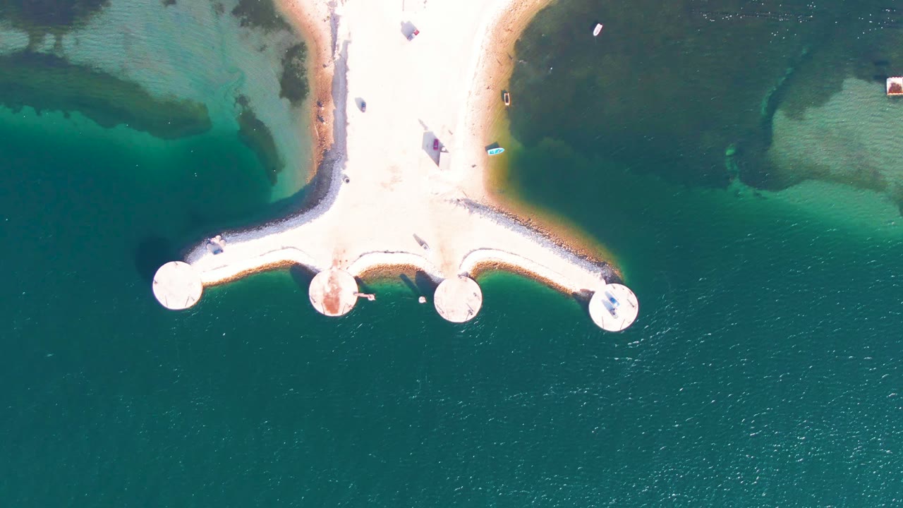 Top-down Guerrero Negro view with bright circular jetty ends, a pale walkway, broad teal water below, and shallow sandy color variations near the shore, with no visible people.