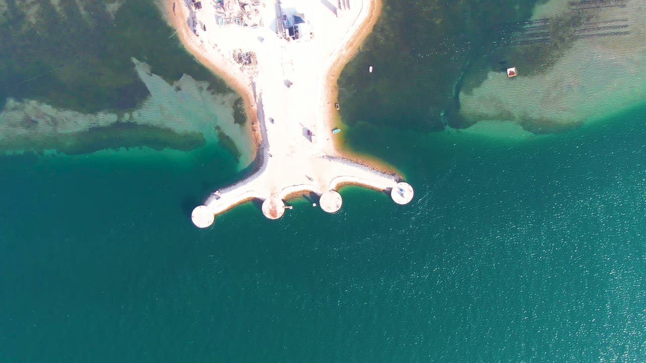 Overhead frame in Guerrero Negro, Baja California Sur showing a pale sandy point with a small dock and round platforms over turquoise water, with no visible people.