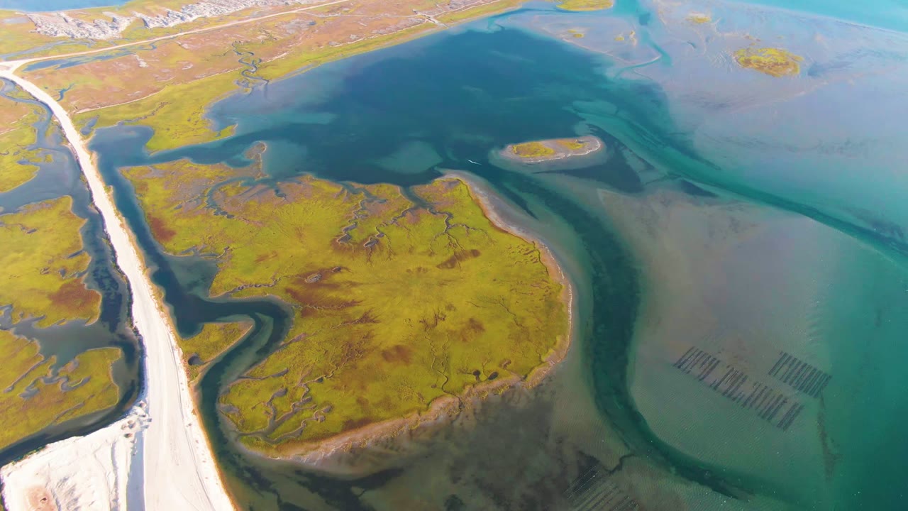 High aerial view over Guerrero Negro wetland terrain with a white road along the left edge, green marsh islands, blue shallow water, and no visible people.
