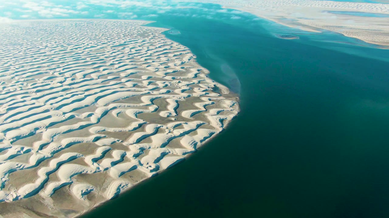 Aerial view over Guerrero Negro dune ridges bordering calm turquoise water, with a small distant sand patch and no visible people.