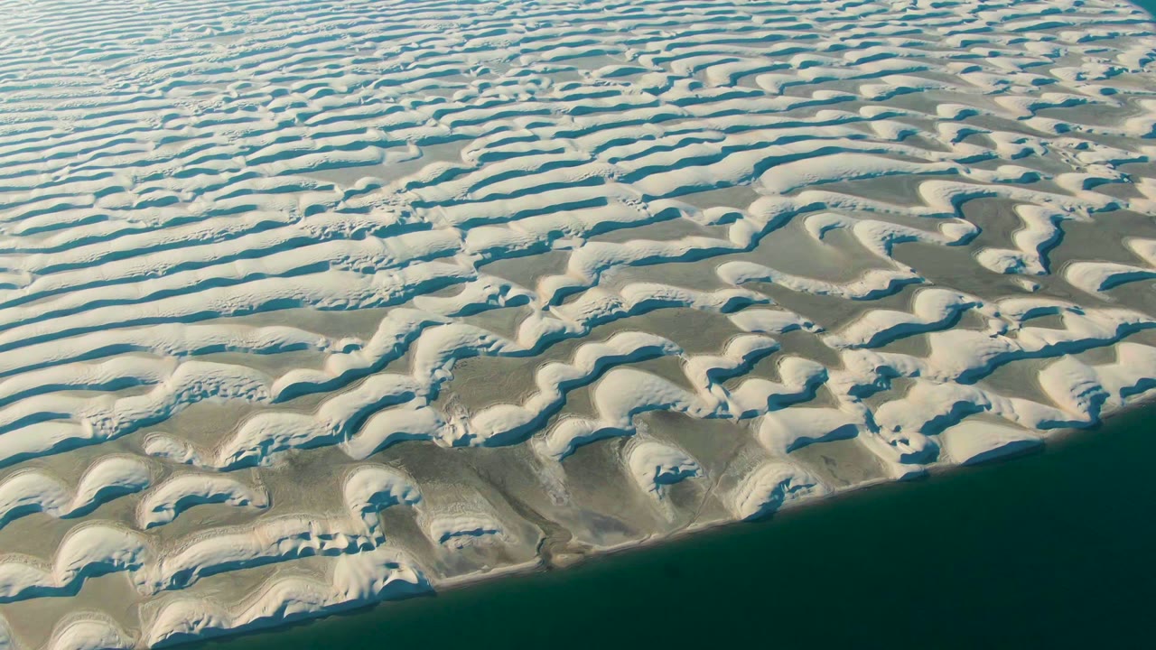 Aerial view over white wave-like sand dunes near the ocean edge in Guerrero Negro, Baja California Sur, with no visible people.