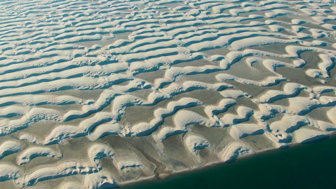 Aerial frame from Guerrero Negro showing rounded white dune forms, blue shadow lines, beige sand pockets, and dark ocean water with no visible people.