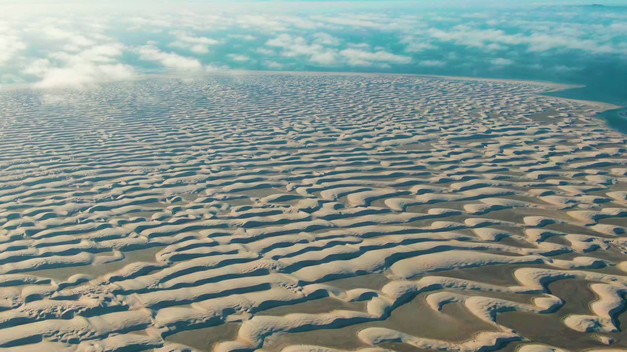 Wide aerial dune field with pale sand ridges and blue-white clouds.