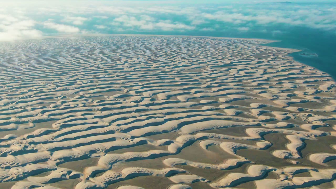 Aerial frame over Guerrero Negro dunes with long rippled ridges filling the lower frame, blue-green water along the right side, and no visible people.