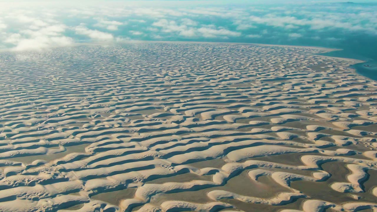Opening frame over Guerrero Negro, Baja California Sur, Mexico, showing light brown wavy sand dunes stretching to a low partly cloudy horizon with no visible people.