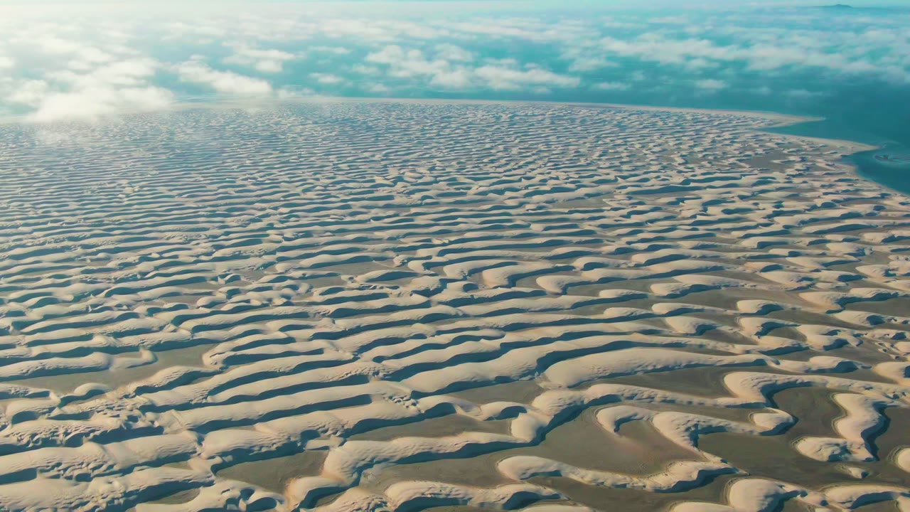 Wide aerial desert view with wavy dune ridges and clouds near the horizon.